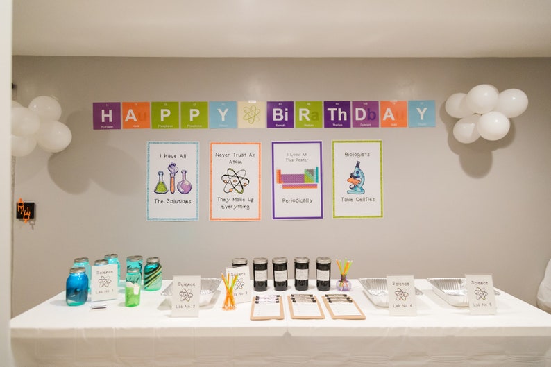 May include: A science-themed birthday party decoration with a banner that reads "HAPPY Bi Ra Th D A Y" using the periodic table elements. There are five posters with science puns and illustrations. The table is decorated with jars of blue and green liquid, jars of black liquid, and signs with the periodic table symbol for science.