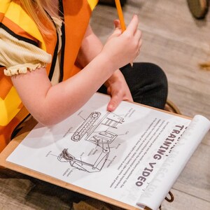 May include: A child wearing an orange and yellow safety vest sits on the floor and looks at a training video on a clipboard. The video shows a diagram of a construction vehicle with labeled parts.