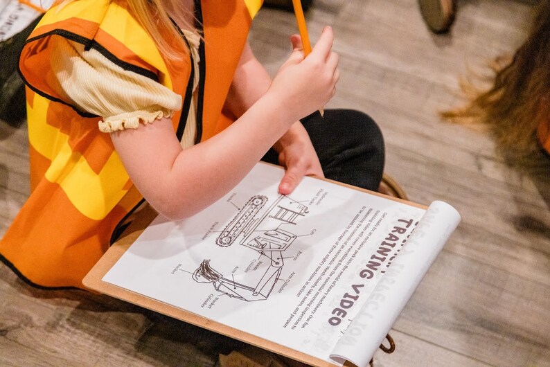 May include: A child wearing an orange and yellow safety vest sits on the floor and looks at a training video on a clipboard. The video shows a diagram of a construction vehicle with labeled parts.