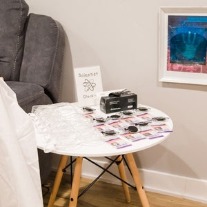 May include: A white table with a black box, clear plastic containers, and small purple cards with black and white images. The table has a white top and wooden legs. A white sign with the text "Scientist Check-in" is on the table. The sign has a black and white drawing of an atom.