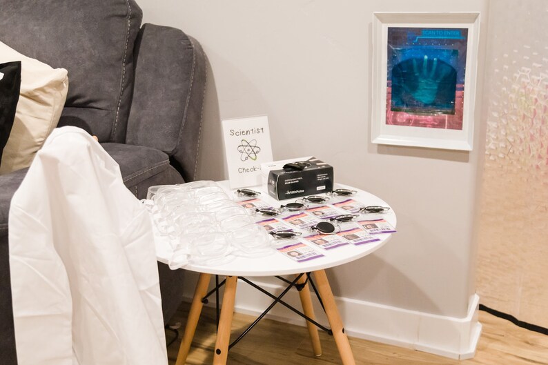 May include: A white table with a black box, clear plastic containers, and small purple cards with black and white images. The table has a white top and wooden legs. A white sign with the text "Scientist Check-in" is on the table. The sign has a black and white drawing of an atom.
