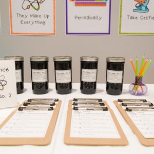May include: A science themed party table with six glass jars labeled "Specimen No. 1" through "Specimen No. 6". The jars are filled with a dark liquid. There are also clipboards with handwritten lists and a sign that says "Science Lab No. 3".