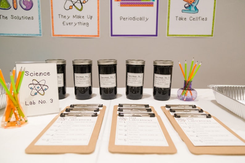 May include: A science themed party table with six glass jars labeled "Specimen No. 1" through "Specimen No. 6". The jars are filled with a dark liquid. There are also clipboards with handwritten lists and a sign that says "Science Lab No. 3".