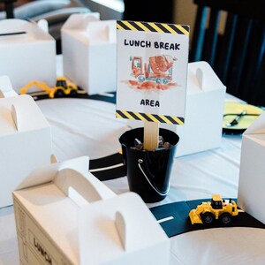 May include: A construction themed party table setting with white lunch boxes, toy construction vehicles, and a sign that reads "Lunch Break Area" with a drawing of a cement mixer truck.