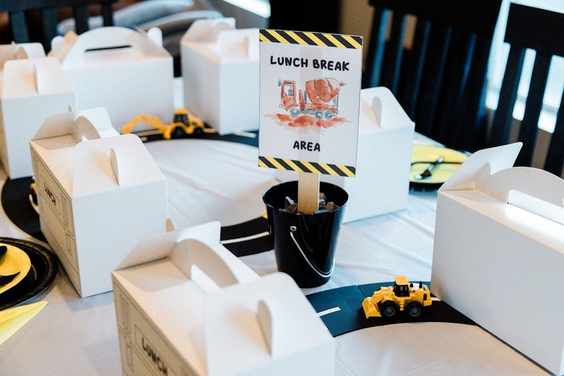 May include: A construction themed party table setting with white lunch boxes, toy construction vehicles, and a sign that reads "Lunch Break Area" with a drawing of a cement mixer truck.