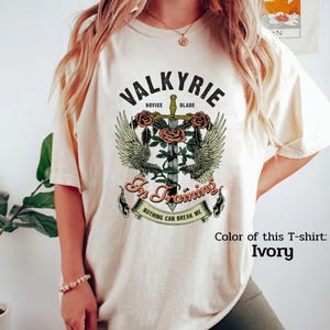 May include: Ivory-colored t-shirt with a graphic design featuring the word "VALKYRIE" above a sword, roses, and wings. The text "In Training" and "Nothing Can Break Me" are also included in the design.