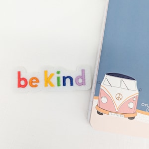 May include: A rainbow-coloured embroidered patch reading "be kind". The patch is on a white surface next to a notebook featuring a pink and orange van illustration and the words "enjoy the ride".