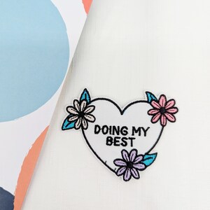 May include: White fabric with an embroidered heart-shaped patch that says "Doing My Best" surrounded by three pink, blue, and white flowers.