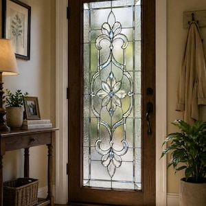 Puede incluir: Una puerta de entrada de madera con una gran ventana de vidrieras ornamentada. El vidrio presenta un diseño floral con detalles claros y azul claro. La puerta está en un recibidor de tonos neutros con una mesa auxiliar, una lámpara y una planta en maceta.