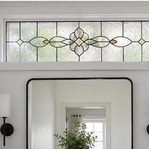 Peut inclure: Une fenêtre imposte rectangulaire dotée d'un cadre blanc et de panneaux en verre texturé. Le centre de la fenêtre présente un motif décoratif en plomb avec un dessin floral symétrique en verre clair et jaune pâle serti de métal noir.