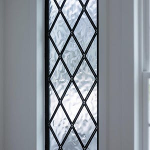 Puede incluir: Una ventana rectangular con un patrón de rejilla de metal negro. El vidrio tiene una apariencia texturizada y ondulada. La ventana está enmarcada en blanco, con un vistazo a otra ventana a la derecha.