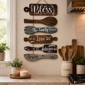 May include: A vertical wall hanging composed of six wooden kitchen utensil-shaped plaques connected by twine. The plaques feature white and black text reading: Bless the FOOD BEFORE US, The Family BESIDE US, and the Love between us Amen.