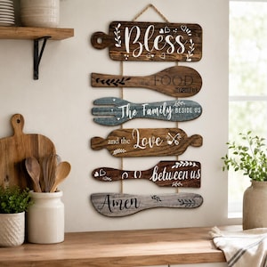 Peut inclure: Une d&eacute;coration murale verticale compos&eacute;e de six plaques en bois en forme d'ustensiles de cuisine reli&eacute;es par une corde. Les plaques portent les inscriptions : Bless, the FOOD BEFORE US, The Family BESIDE US, and the Love, between us, et Amen.