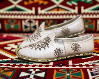 May include: A pair of white leather slippers with intricate brown geometric designs. The slippers are on a colorful rug with a traditional pattern.