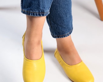 May include: A pair of bright yellow leather ballet flats with a pointed toe. The flats are being worn by someone wearing blue jeans.