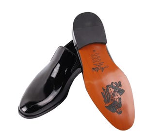 May include: Pair of glossy black leather slip-on shoes. The soles are brown with a black graphic design and the brand name "Antao". The shoes are size 42.
