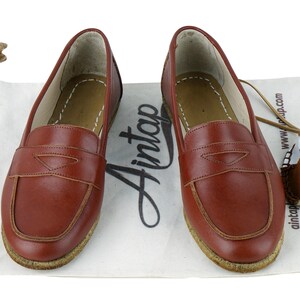 May include: A pair of brown leather loafers with a suede sole. The loafers have a simple design with a single strap across the top. The loafers are sitting on a white fabric surface.