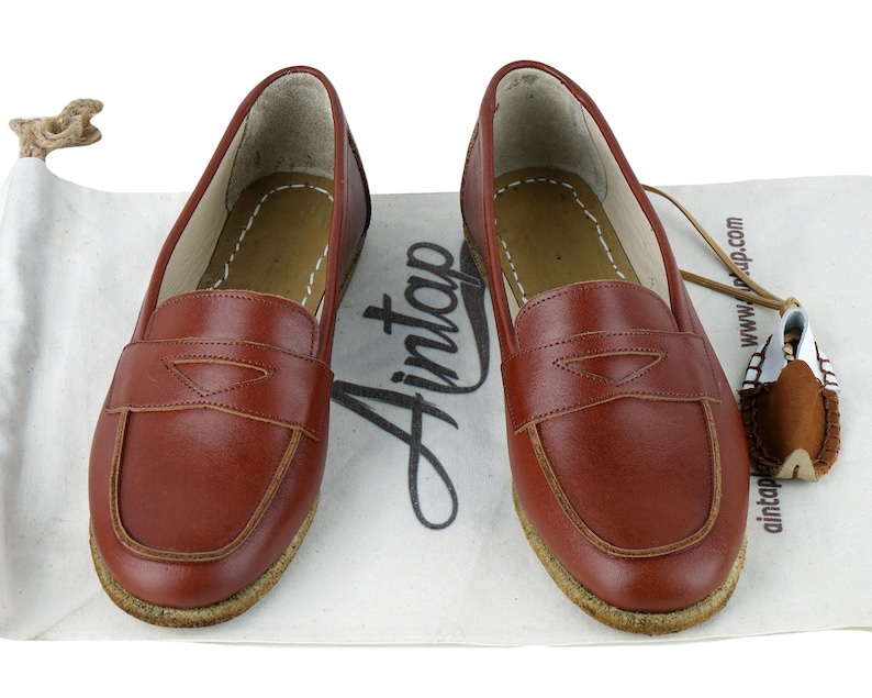 May include: A pair of brown leather loafers with a suede sole. The loafers have a simple design with a single strap across the top. The loafers are sitting on a white fabric surface.