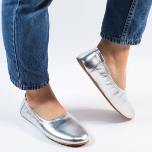 Puede incluir: Un par de bailarinas de cuero plateado con punta redonda. Las bailarinas las lleva puestas una persona que lleva pantalones vaqueros azules.