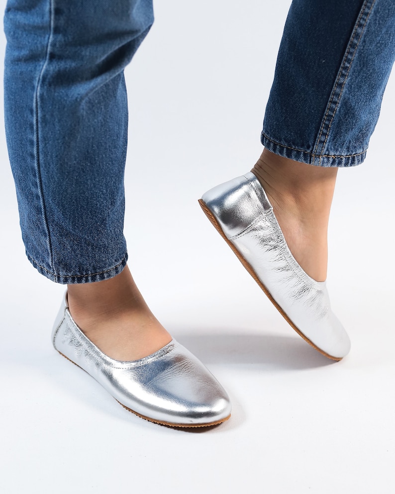 Puede incluir: Un par de bailarinas de cuero plateado con punta redonda. Las bailarinas las lleva puestas una persona que lleva pantalones vaqueros azules.