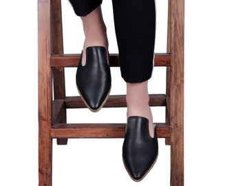 May include: A pair of black leather pointed-toe mules with a low heel. The shoes are worn by a person sitting on a wooden stool.