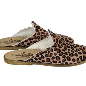 Op de afbeelding: Een paar bruine en zwarte pantoffels met luipaardprint en een witte voering van bont. De pantoffels zijn gemaakt van leer en hebben een bruine leren zool. De pantoffels hebben de tekst "Antap" op de zool.