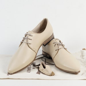 May include: Two pairs of beige leather lace-up shoes with pointed toes. The shoes are on a white surface with a brown fabric bag.