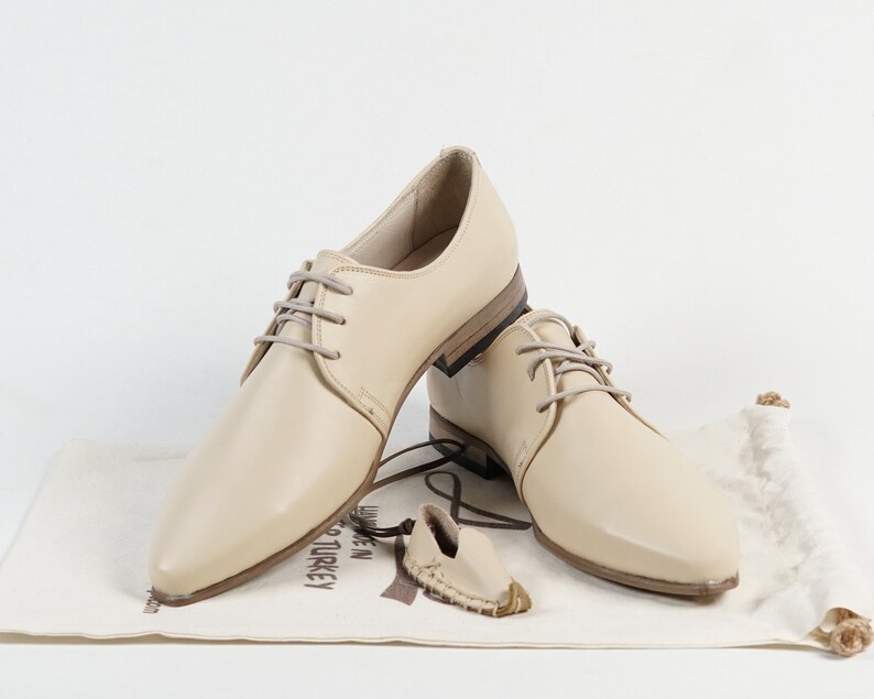 May include: Two pairs of beige leather lace-up shoes with pointed toes. The shoes are on a white surface with a brown fabric bag.