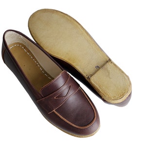 May include: A pair of brown leather loafers with a brown leather sole. The loafers have a simple design with a single strap across the top.