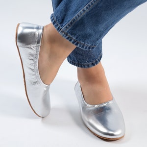 Puede incluir: Un par de bailarinas de cuero plateado con punta redonda. Las bailarinas las lleva una persona que lleva pantalones vaqueros azules.