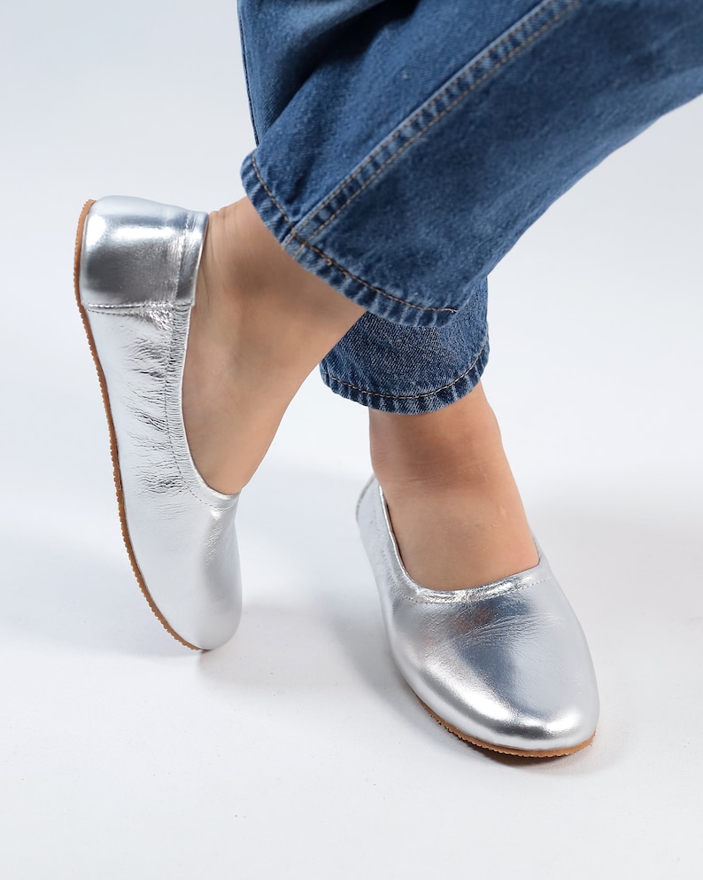 Puede incluir: Un par de bailarinas de cuero plateado con punta redonda. Las bailarinas las lleva una persona que lleva pantalones vaqueros azules.