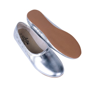 Puede incluir: Un par de bailarinas de cuero plateado con suela de goma marr&oacute;n. Las bailarinas tienen un dise&ntilde;o sencillo y est&aacute;n hechas de un material suave y c&oacute;modo.