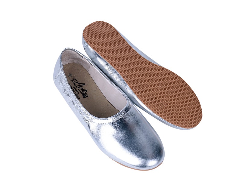 Puede incluir: Un par de bailarinas de cuero plateado con suela de goma marr&oacute;n. Las bailarinas tienen un dise&ntilde;o sencillo y est&aacute;n hechas de un material suave y c&oacute;modo.