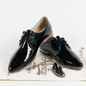 May include: Two pairs of black patent leather lace-up shoes with a pointed toe. The shoes are on a white surface with a small brown bag and a brown tag that says "Lintop".