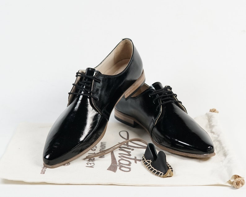 May include: Two pairs of black patent leather lace-up shoes with a pointed toe. The shoes are on a white surface with a small brown bag and a brown tag that says "Lintop".