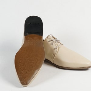 May include: A pair of cream-colored leather shoes with a pointed toe and lace-up closure. The shoes have a brown leather sole with a textured pattern.