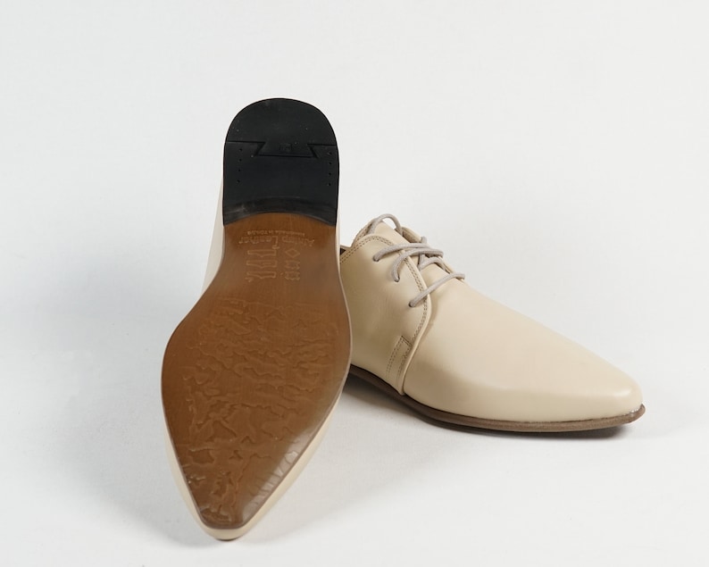 May include: A pair of cream-colored leather shoes with a pointed toe and lace-up closure. The shoes have a brown leather sole with a textured pattern.