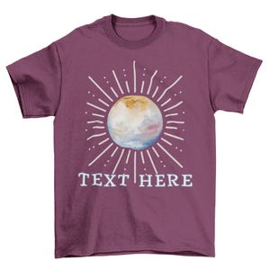 May include: A maroon t-shirt featuring a watercolor design of a sun with radiating lines and the text "TEXT HERE" in white. The sun design has shades of yellow, blue, and white.