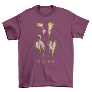 May include: Burgundy t-shirt featuring a watercolor illustration of Venus flytraps in shades of green, pink, and white. The text "TEXT HERE" is printed below the illustration. The shirt has a crew neck and short sleeves.