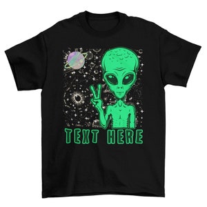 May include: Black t-shirt with a green alien graphic. The alien is making a peace sign and is surrounded by stars. The text "TEXT HERE" is printed below the alien.