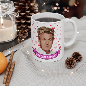 May include: White ceramic coffee mug with a cartoon image of Gordon Ramsay and the text "Gordon Ramsay" in a pink banner. The mug is decorated with pink hearts.