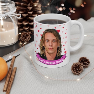 May include: White ceramic mug with a pink heart pattern and a photo of Jamie Campbell Bower. The mug has a pink ribbon with the text "Jamie Campbell Bower" on it.