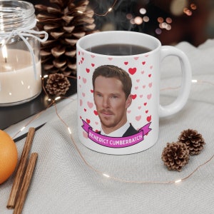 May include: White ceramic mug with a pink heart pattern and a cartoon image of Benedict Cumberbatch. The mug has a pink ribbon with the text "Benedict Cumberbatch" on it.