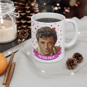 May include: White ceramic mug with a pink heart pattern and a cartoon image of Peter Falk. The mug has a pink ribbon with the text "Peter Falk" in white letters.