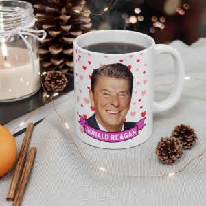 May include: White ceramic coffee mug with a cartoon image of Ronald Reagan and pink hearts. The mug has the text "RONALD REAGAN" in a pink banner.