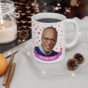 May include: White ceramic mug with a cartoon image of Lester Holt, a news anchor, with a pink ribbon that says "Lester Holt" and a background of pink hearts.
