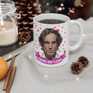 May include: White ceramic mug featuring a portrait of Daniel Day-Lewis, surrounded by pink and red hearts. The mug has a handle and the name "DANIEL DAY-LEWIS" is printed on a pink banner below the portrait. The mug is filled with a dark liquid.