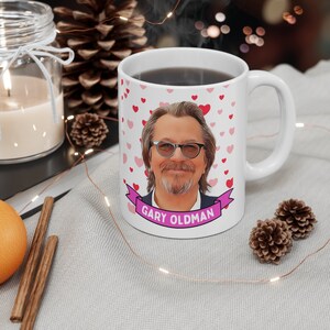 Puede incluir: Taza de cerámica blanca con un patrón de corazones rosas y una imagen de dibujos animados de Gary Oldman. La taza tiene una cinta rosa con el texto "Gary Oldman" en ella.