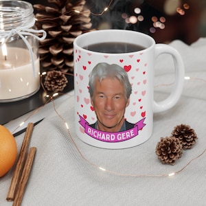 May include: White ceramic mug with a pink heart pattern and a photo of Richard Gere. The mug has a pink ribbon with the text "Richard Gere" on it.