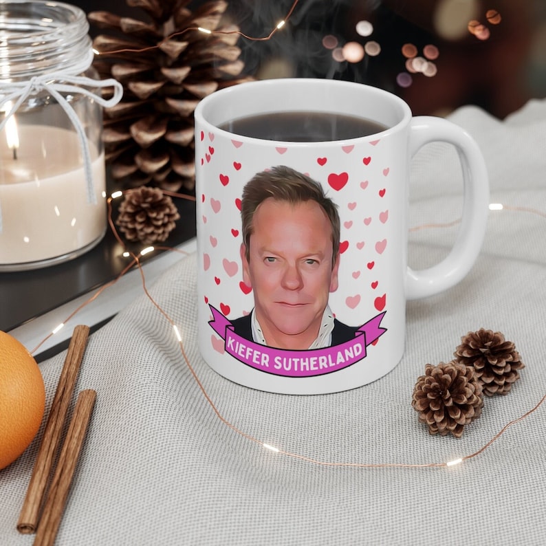 May include: White ceramic mug with a pink ribbon and the text "KIEFER SUTHERLAND". The mug features a color image of Kiefer Sutherland with a background of pink and red hearts.